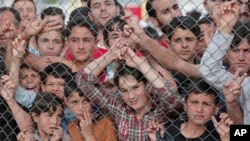 Para migran berdiri di belakang pagar kamp pengungsi Nizip di provinsi Gaziantep, Turki bagian tenggara (23/4). (AP/Lefteris Pitarakis)
