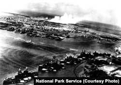 This image was captured by a Japanese naval aviator in the opening moments of the attack on Pearl Harbor. Torpedo tracks can be seen headed towards Battleship Row. Smoke rises in the distance from the burning aircraft and hangars at Hickam Field.